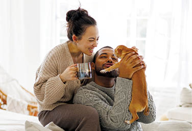 Smiling asian woman in sweater holding coffee near african american boyfriend playing with dog at home